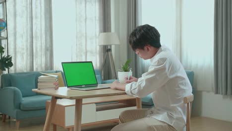 side view of asian man student look at green screen computer and write in the notebook while studying online at home