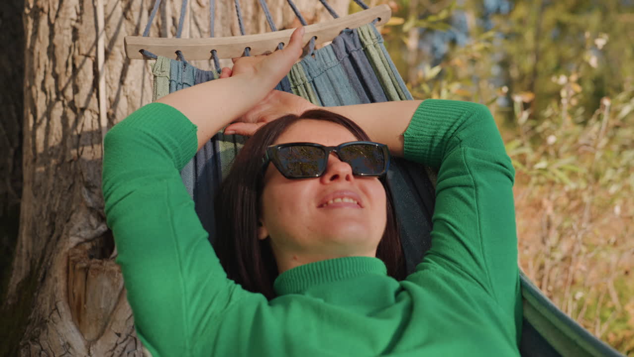 Mujer en hamaca durante un campamento, recostada bajo un tronco de árbol con gafas de sol, suéter verde, luz solar moteada en la pradera, sonriendo y soñando despierta, descansando entre caminatas como escritora de viajes