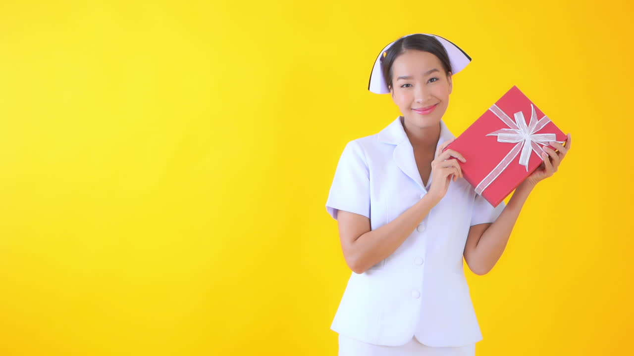 Happy Asian nurse holding a gift box in her hands and smiling posing isolated on a yellow background, slow-motion