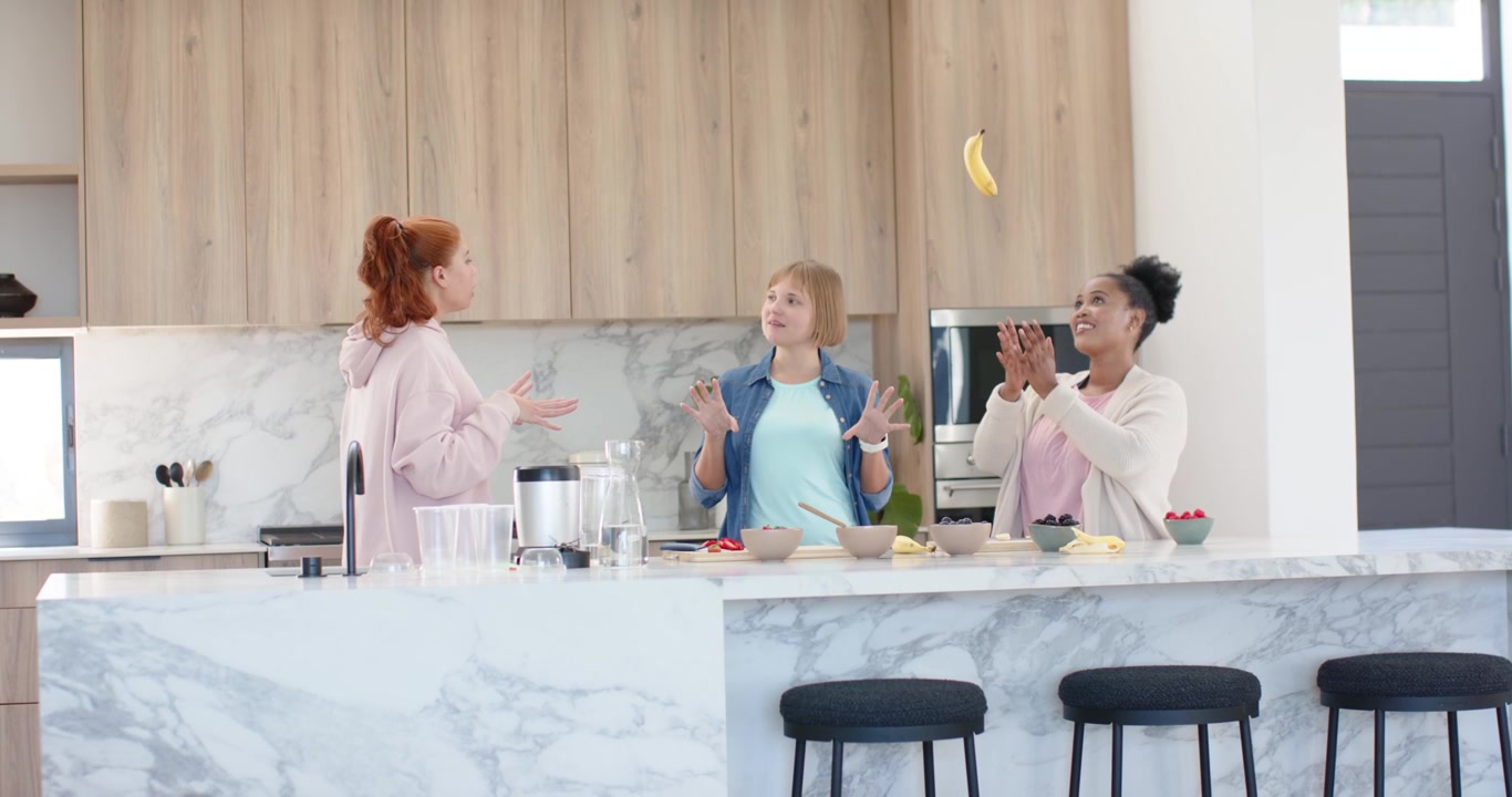 Donne di diverse etnie che chiacchierano mentre preparano la frutta su un'isola di marmo in cucina, lanciando una banana per ridere.