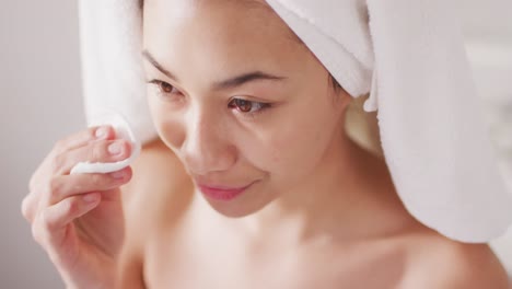 Video-of-portrait-of-smiling-biracial-woman-with-towel-on-hair-cleansing-face-in-bathroom