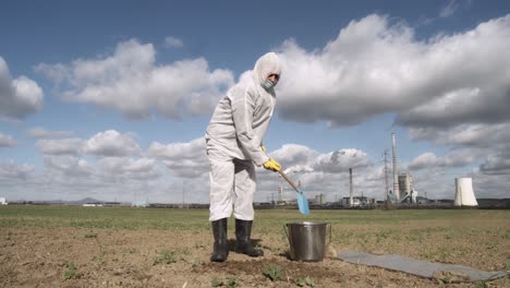 muestreo de suelo después de un desastre ecológico, científico cavando terreno de campo con fábrica en segundo plano