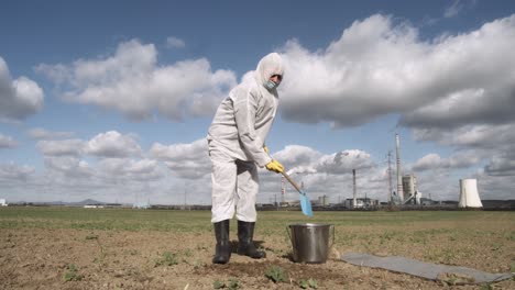 soil sampling after ecological disaster, scientist digging field ground with factory in background