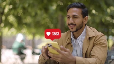 hombre sonriente sentado en una mesa al aire libre mirando un teléfono móvil con emojis de gráficos de movimiento que muestran múltiples notificaciones de redes sociales o gustos que reaccionan al contenido en línea