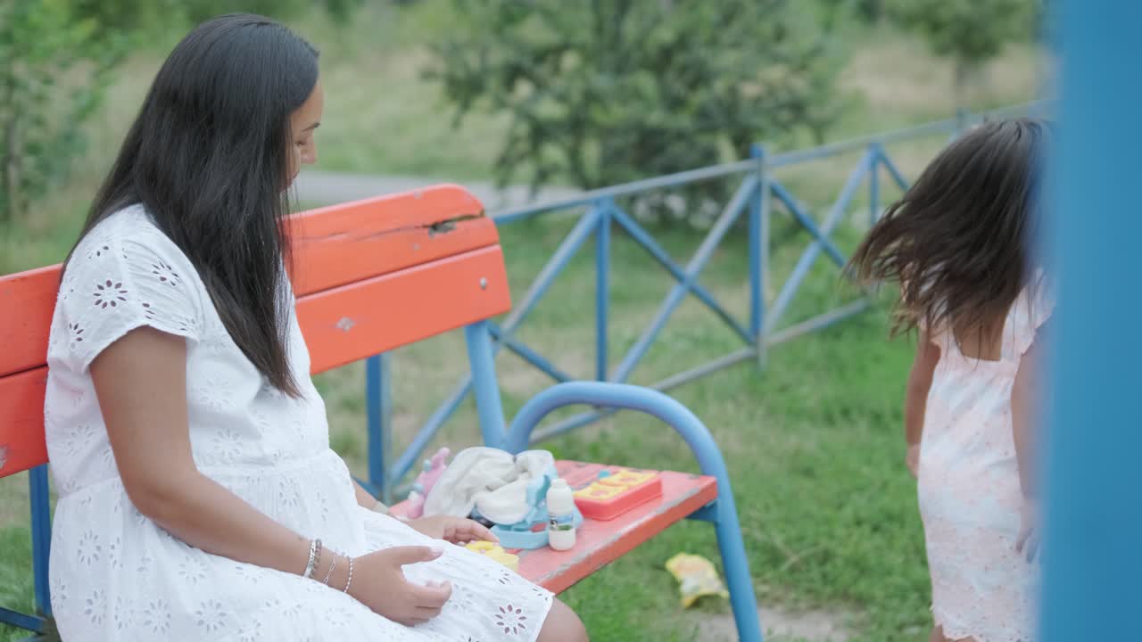 임신 한 어머니 와 어린 딸 은 나무 와 초록색 으로 둘러싸인 공원 의 놀이터 에서 함께 놀이 를 즐긴다