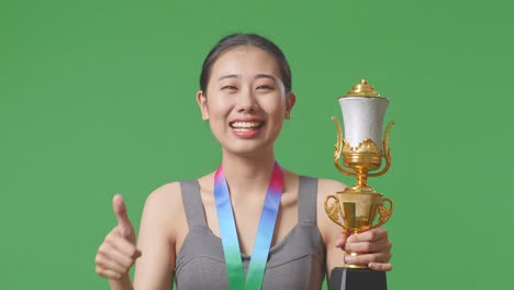 close up of asian woman with a gold medal and trophy showing thumbs up gesture and smiling to camera as the first winner on green screen background in the studio