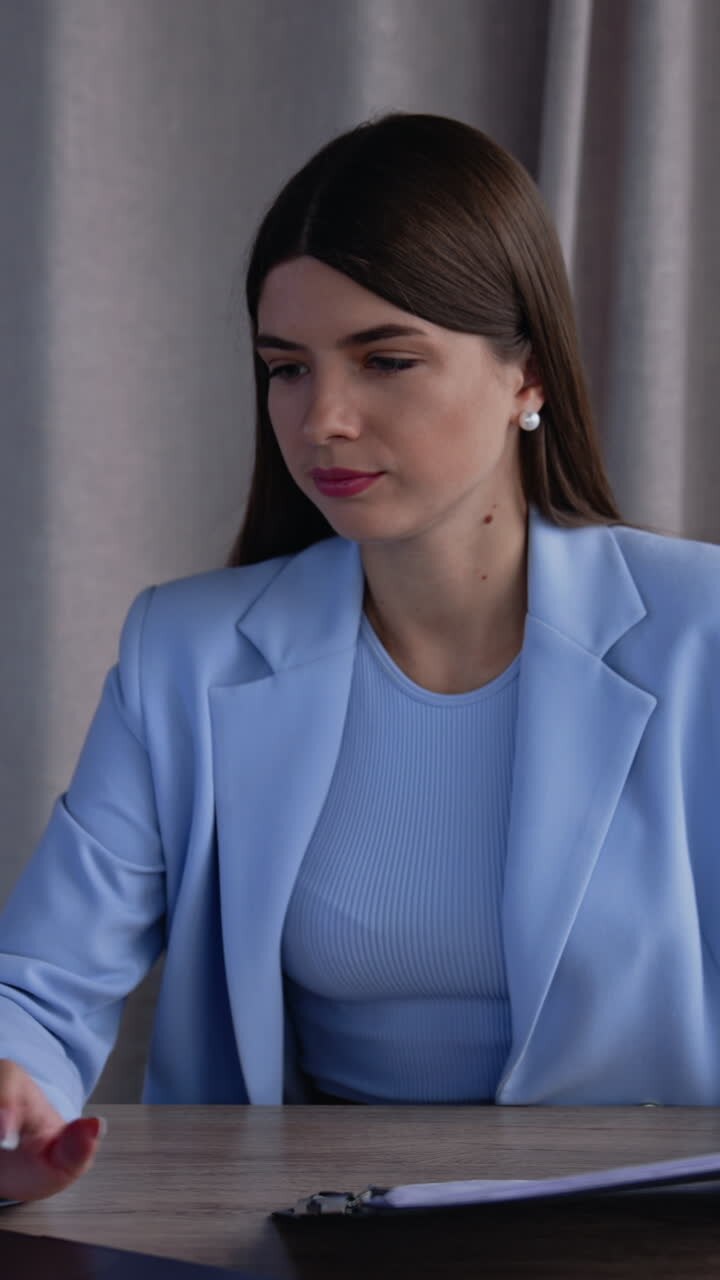 Dark-haired young woman works at laptop in office. An at the next table talks and laughs to a female blonde colleague standing beside him. Vertical video