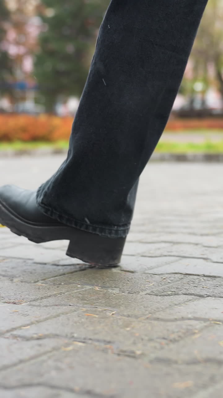 Side view of person's legs in black trousers and black boots walking slowly on outdoor paved path during calm overcast day, capturing subtle foot movement with soft background of trees