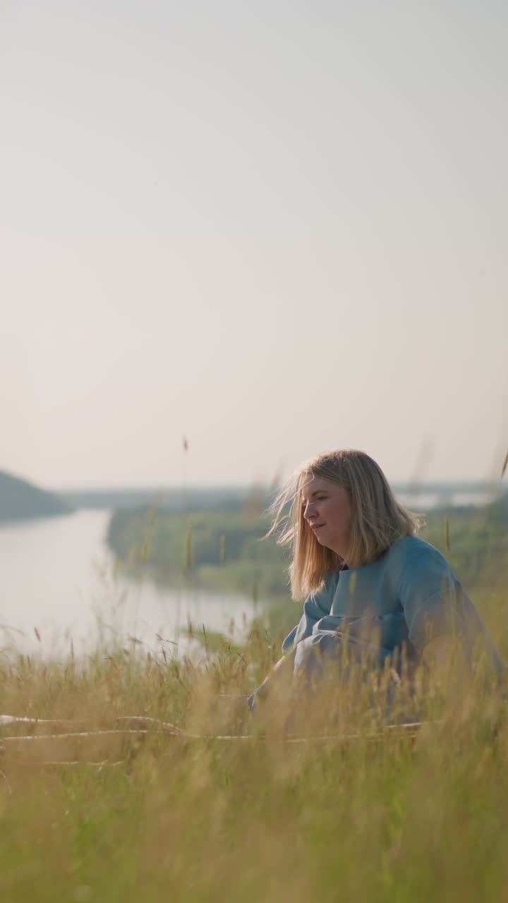 A woman in a blue gown and her sons in white shirts hold a large plaid scarf on a breezy hilltop beside a scenic lake. The family enjoys the fresh air and the natural beauty around them