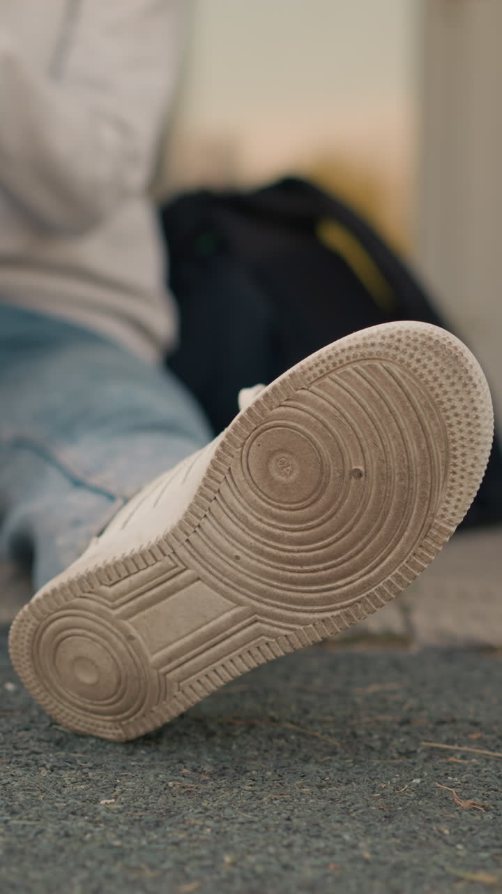 primer plano de la pierna del individuo sentado, centrándose en la suela de la zapatilla blanca, con la mano descansando en la pierna y la bolsa negra cerca, el entorno al aire libre casual hace hincapié en el estado de ánimo relajado y relajado