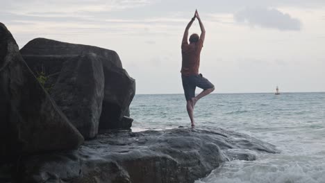 Adult-Man-Trying-To-Do-A-Yoga-Pose-By-Standing-On-One-Leg-In-The-Shoreline-With-Crashing-Waves