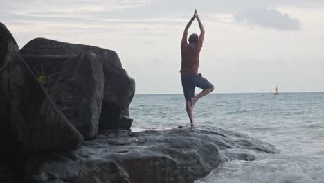 Hombre-Adulto-Tratando-De-Hacer-Una-Pose-De-Yoga-Parándose-Sobre-Una-Pierna-En-La-Costa-Con-Olas-Rompiendo