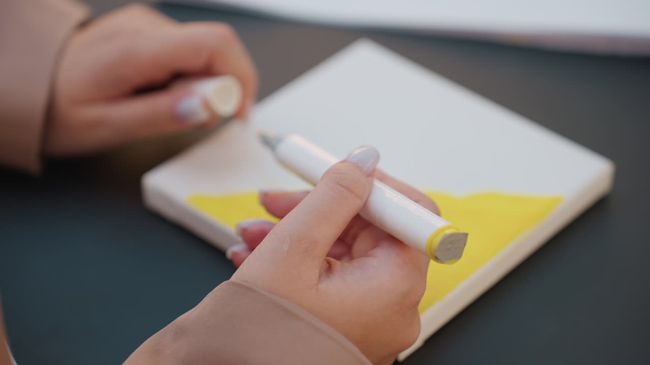 Un artista examina el equipo; los detalles precisos requieren una configuración cuidadosa, la preparación de las herramientas de forma precisa para un trabajo de ilustración detallado y las manos retiran cuidadosamente el tapón del rotulador sobre un material amarillo vibrante.