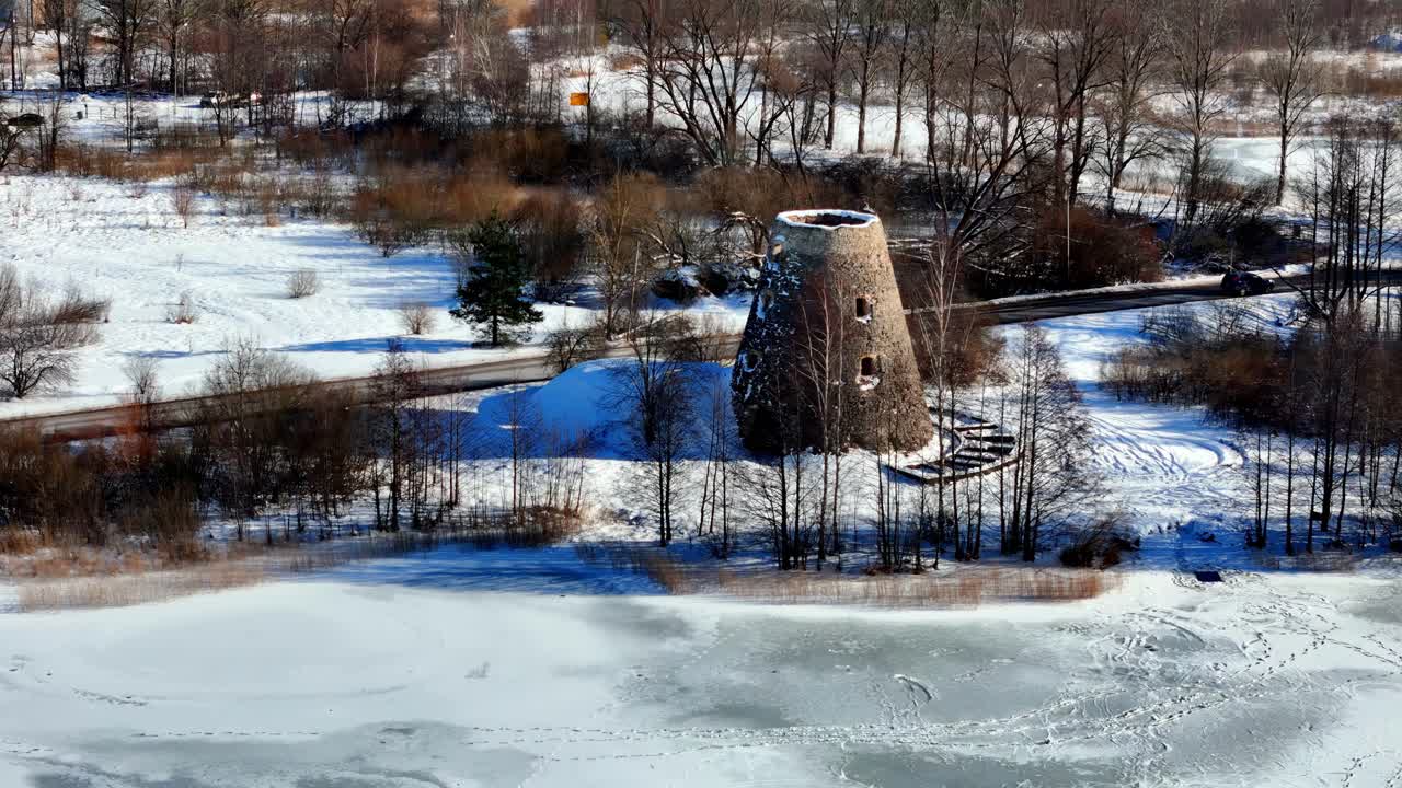 A weathered stone tower stands near a partially frozen lake and quiet snow-covered road in the countryside, framed by bare winter trees and distant buildings, seen from above.