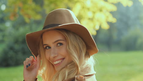 vista de cerca de una joven rubia sosteniendo su sombrero y sonriendo a la cámara en el parque en otoño