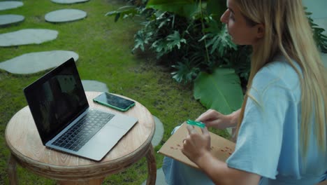 una chica en un vestido azul remotamente en línea trabajando detrás de una computadora portátil y mirando a la pantalla el patio trasero con plantas verdes