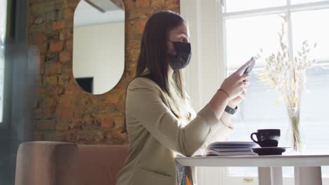 caucasian female customer wearing face mask, sitting at table, using smartphone