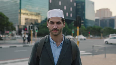 Primer-Plano-Retrato-De-Un-Hombre-Atractivo-Y-Seguro-Del-Medio-Oriente-Que-Mira-Serio-A-La-Cámara-Pensativo-En-El-Fondo-Urbano-De-La-Ciudad-Con-Sombrero-Kufi