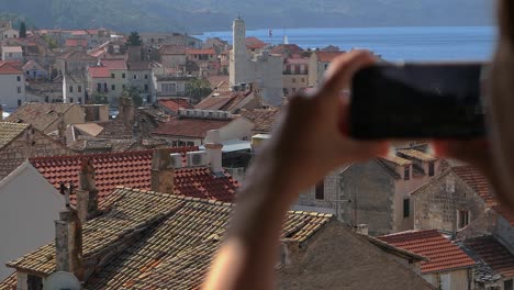 Tourist-taking-photo-of-coastal-old-town-from-above,-Komiza,-Vis-Island,-Croatia