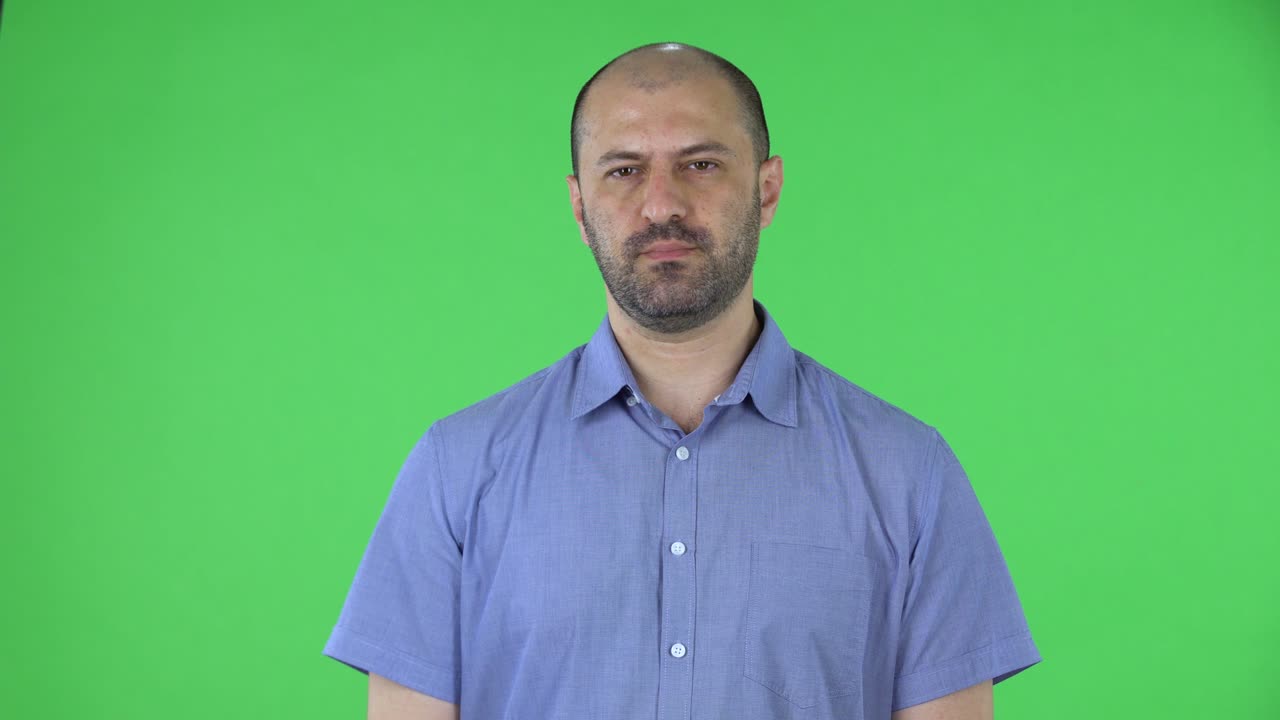 Portrait of a middle aged men looking at the camera. Balding male with a beard in a blue shirt posing on a green screen in the studio. Close up