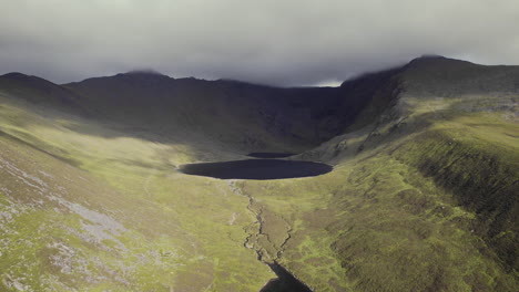 Beautiful-aerial-of-a-small-river-with-a-lough-within-grass-covered-mountains-with-a-stoney-cloud-covered-mountain-range-laying-behind-3