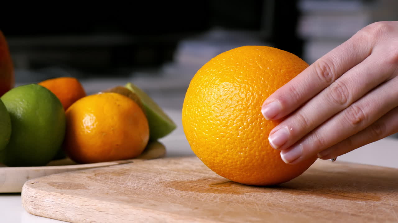 Woman slicing orange in two parts on a wooden board. Fruits on the background. Slow motion