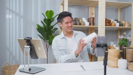 asian handsome man sell vase product online live streaming at home. young business male using laptop computer shows goods to customer and present detail. remote buying and purchase shopping concept.