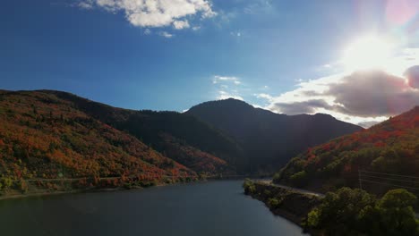 beautiful slow backing up drone shot over a body of water lake reservoir in late autumn fall october november red, orange, and yellow trees covering mountains, blue sky, heavenly sunshine