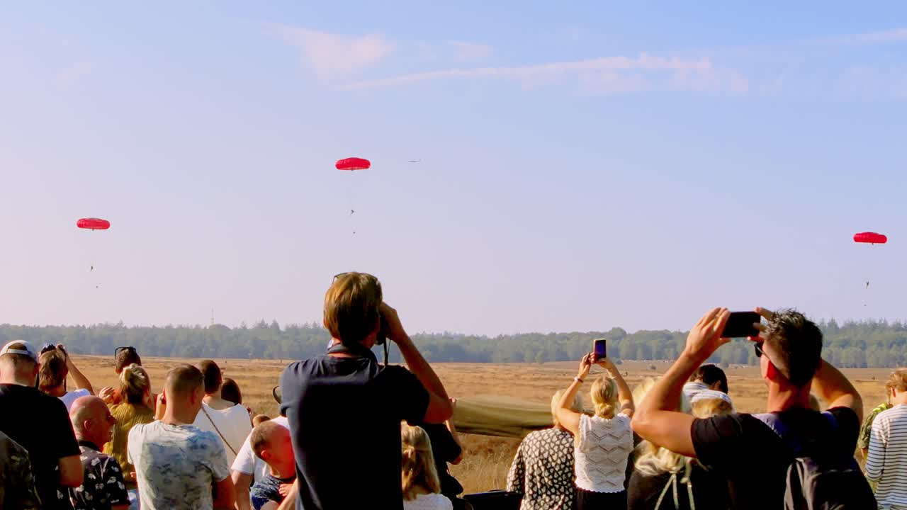 Large crowd watches parachutists with vibrant red canopies descending in the distance during a military event at Ginkelse Heide, Netherlands. Spectators capture the moment on cameras and binoculars