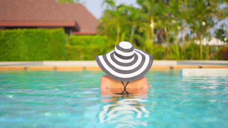 Vista-Trasera-De-Una-Mujer-Con-Un-Gran-Sombrero-A-Rayas-Bañándose-En-Una-Lujosa-Piscina