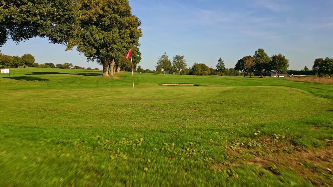Campo de golf verde con búnker de arena y calle recortada bajo un cielo despejado; toma aérea baja que se aleja de la bandera