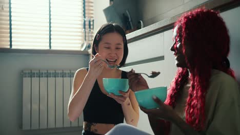 two beautiful diverse friends eat breakfast from a bowl sitting on the floor in the kitchen