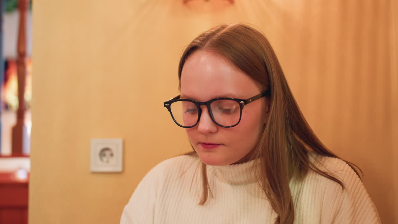 focused lady in white sweater eats food, nodding head with subtle satisfaction, intense expression on face, seated against yellow wall, animated fireplace flame softly glowing from television behind