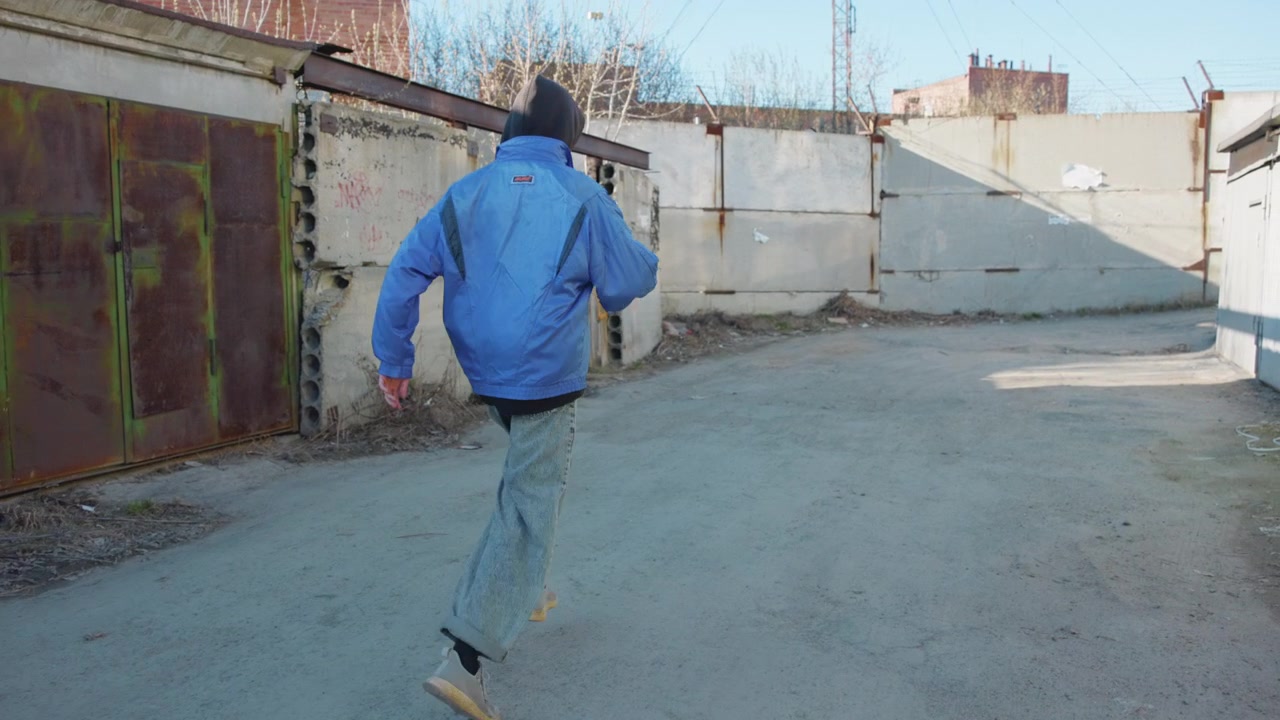 Una persona di spalle con giacca blu si allontana su cemento crepato in un cortile industriale accanto a container metallici, mostrando un tranquillo movimento urbano, superfici usurate, spazio aperto e luce diurna soffusa