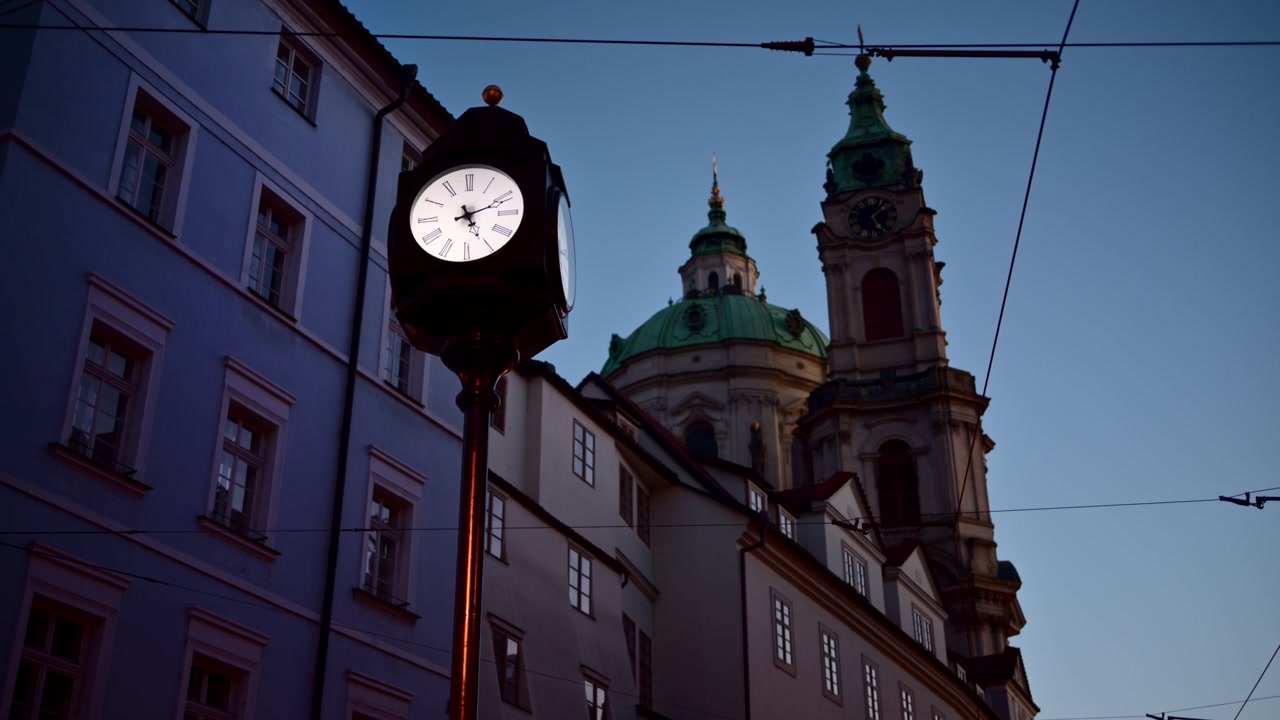 Un orologio stradale illuminato al crepuscolo con la cupola verde e il campanile della Chiesa di San Nicola sullo sfondo, Malá Strana, Praga, Repubblica Ceca