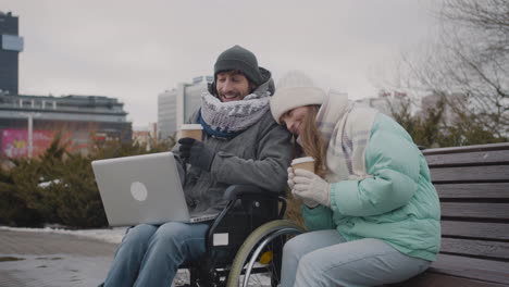 hombre discapacitado en silla de ruedas y su amigo viendo algo divertido en una computadora portátil y riéndose juntos en el parque urbano en invierno 1