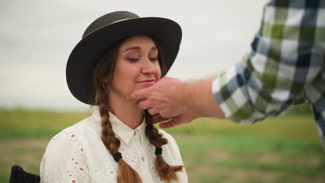 a charming scene where a woman in a white dress and black hat sits on a chair in a serene field. she smiles shyly as a sculptor's hand gently adjusts her hat and hair