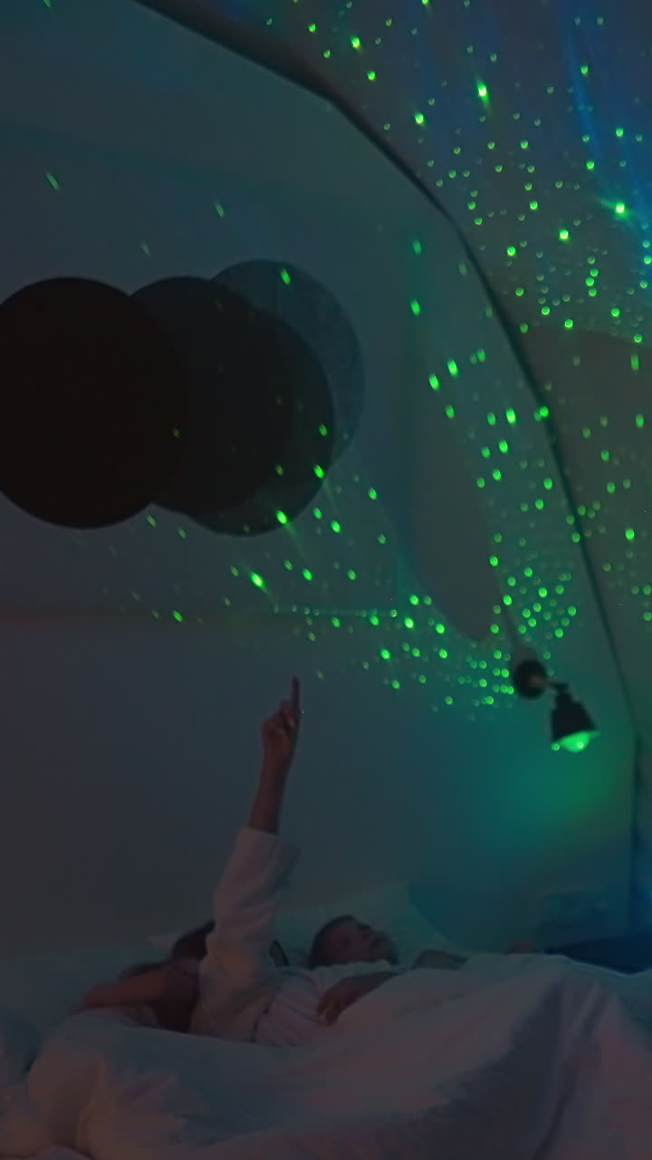 los niños se acuestan en la cama con la madre mirando los rayos de luz nocturna en el techo. la mujer joven señala con la mano los brillantes reflejos de luz nocturada en la habitación