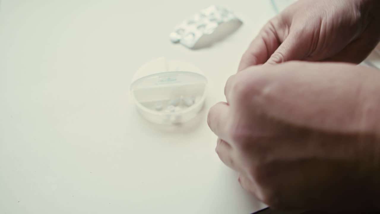 Close up of person placing tablets into weekly pill organizer at home. Suitable for chronic illness management, senior care, and daily health routine themes