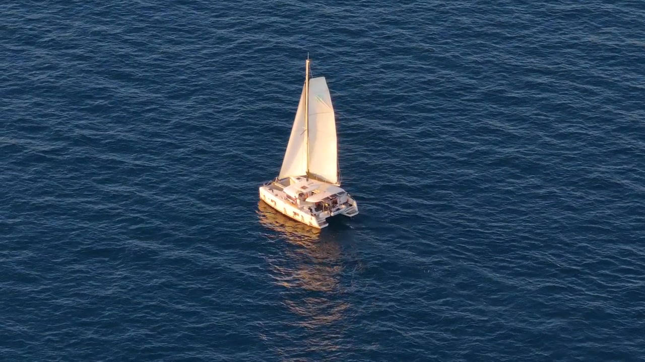 Een boot vaart op open water. De zeilen vangen zonlicht op. De scène toont kalme golven. Geen andere boten zijn zichtbaar. De locatie is waarschijnlijk nabij een kustgebied.