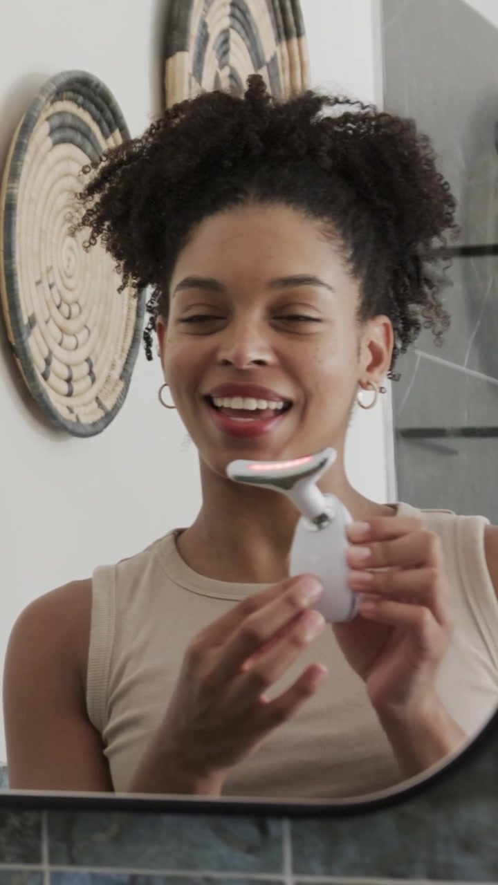 Vertical video: Using woman in beige top showing white device at vanity mirror, turning on red LED