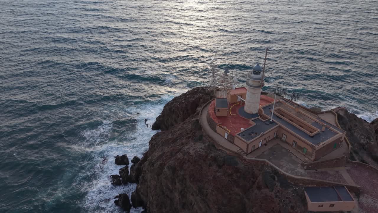 Prospettiva aerea cinematografica dell'iconico faro di Cabo de Gata arroccato su una scogliera spettacolare con onde che si infrangono contro le rocce.