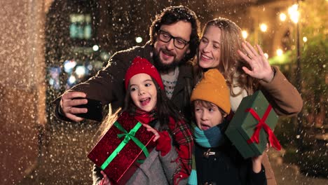 vista de cerca de una familia feliz caucásica sosteniendo regalos y haciéndose un selfie mientras nieva en navidad