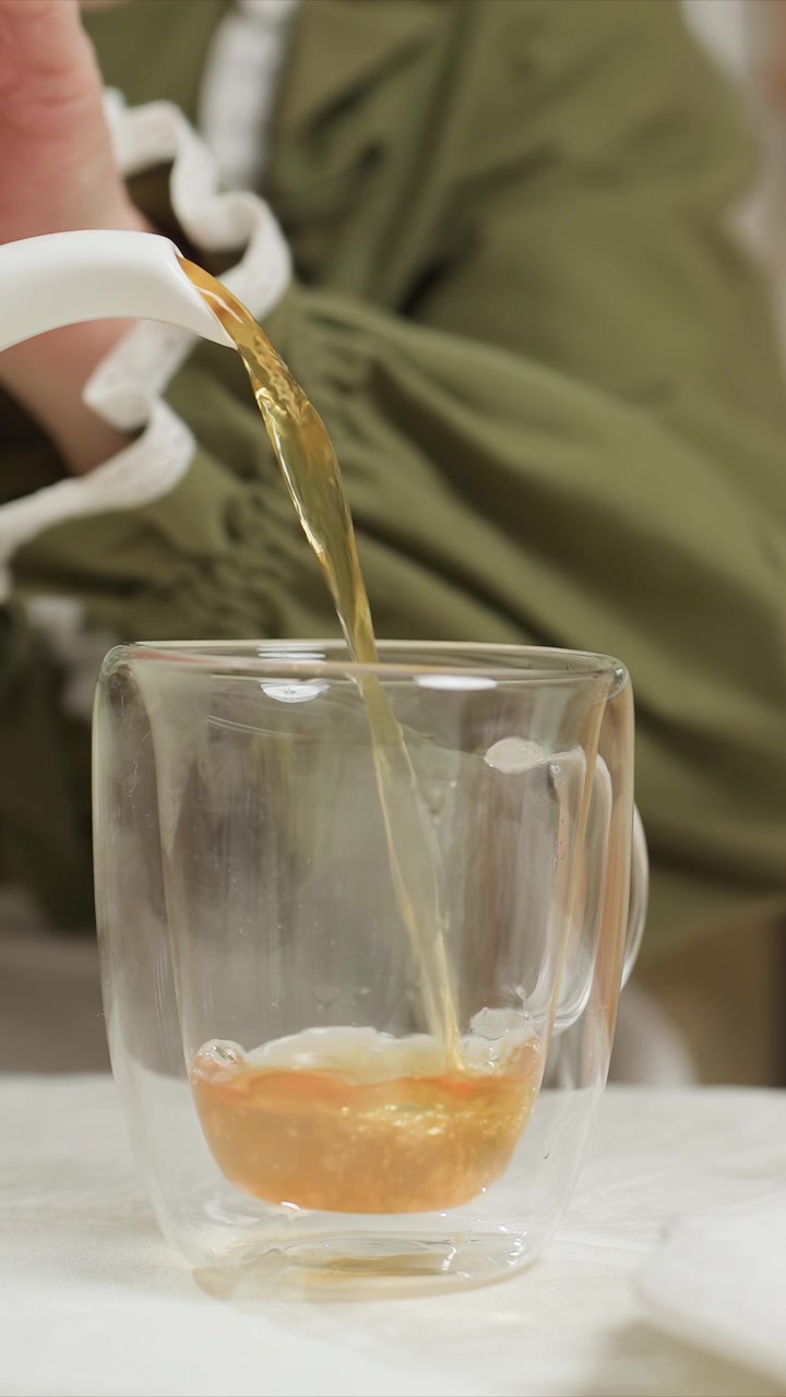 Giovane donna in camicetta verde versa un liquido dorato. Il versamento continuo riempie la tazza costantemente da meno della metà. Una teiera bianca viene usata per versare durante tutta la sequenza, con un'attenzione ravvicinata.