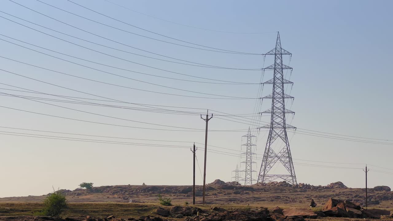 Tall power transmission towers stand across a hazy landscape, with long electric lines stretching into the soft morning sky above distant terrain