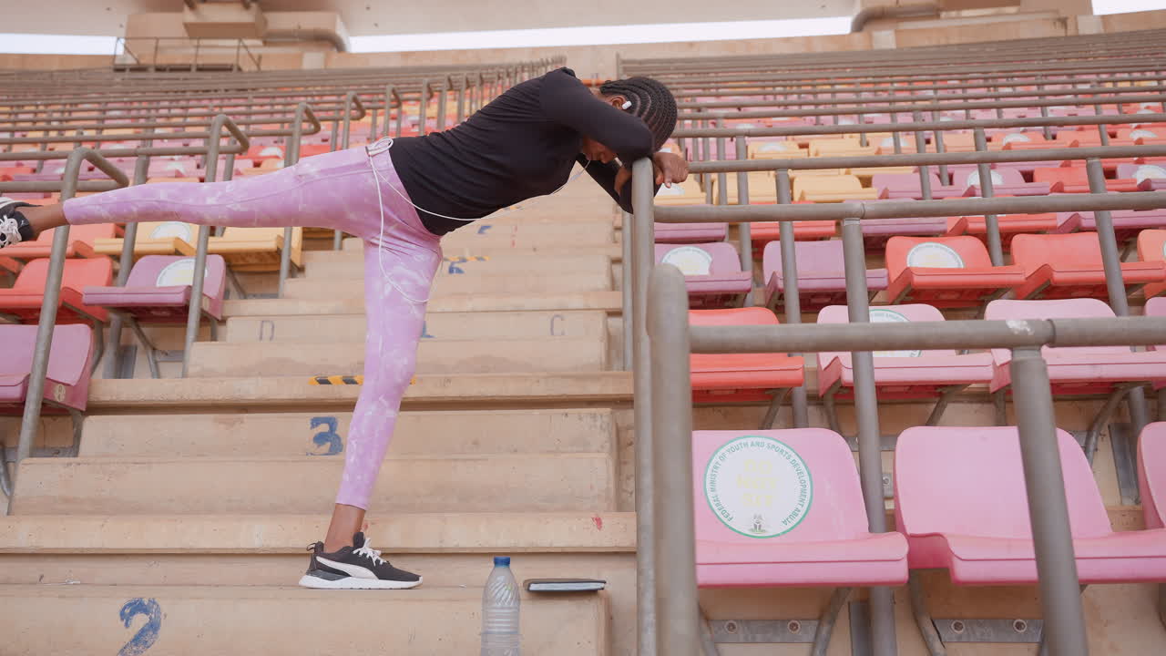 black woman athlete balancing on railing, stadium trainer stretching leg on empty bleachers in pink leggings and sneakers, intense warmup, focused posture, sunrise light, perseverance and recovery