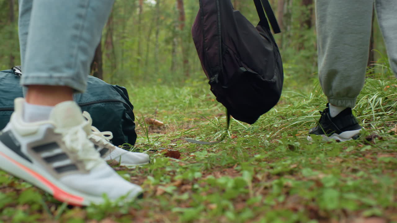 Leg view of two people wearing sneakers, standing still on forest floor after walk, one in joggers and other in jeans, surrounded by greenery, appearing to rest or prepare for break in woodland