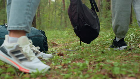 leg view of two people wearing sneakers, standing still on forest floor after walk, one in joggers and other in jeans, surrounded by greenery, appearing to rest or prepare for break in woodland