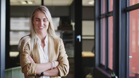 Mujer-De-Negocios-Rubia-Blanca-Milenaria-Sonriendo-A-La-Cámara-Junto-A-La-Ventana-En-Una-Oficina,-De-Cerca