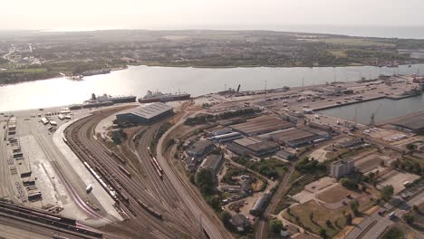 In-this-drone-shot,-a-bustling-port-with-multiple-ships-and-cranes-is-set-by-a-large-water-body,-framed-by-roads,-buildings,-and-a-distant-bridge,-all-under-a-clear-daytime-sky